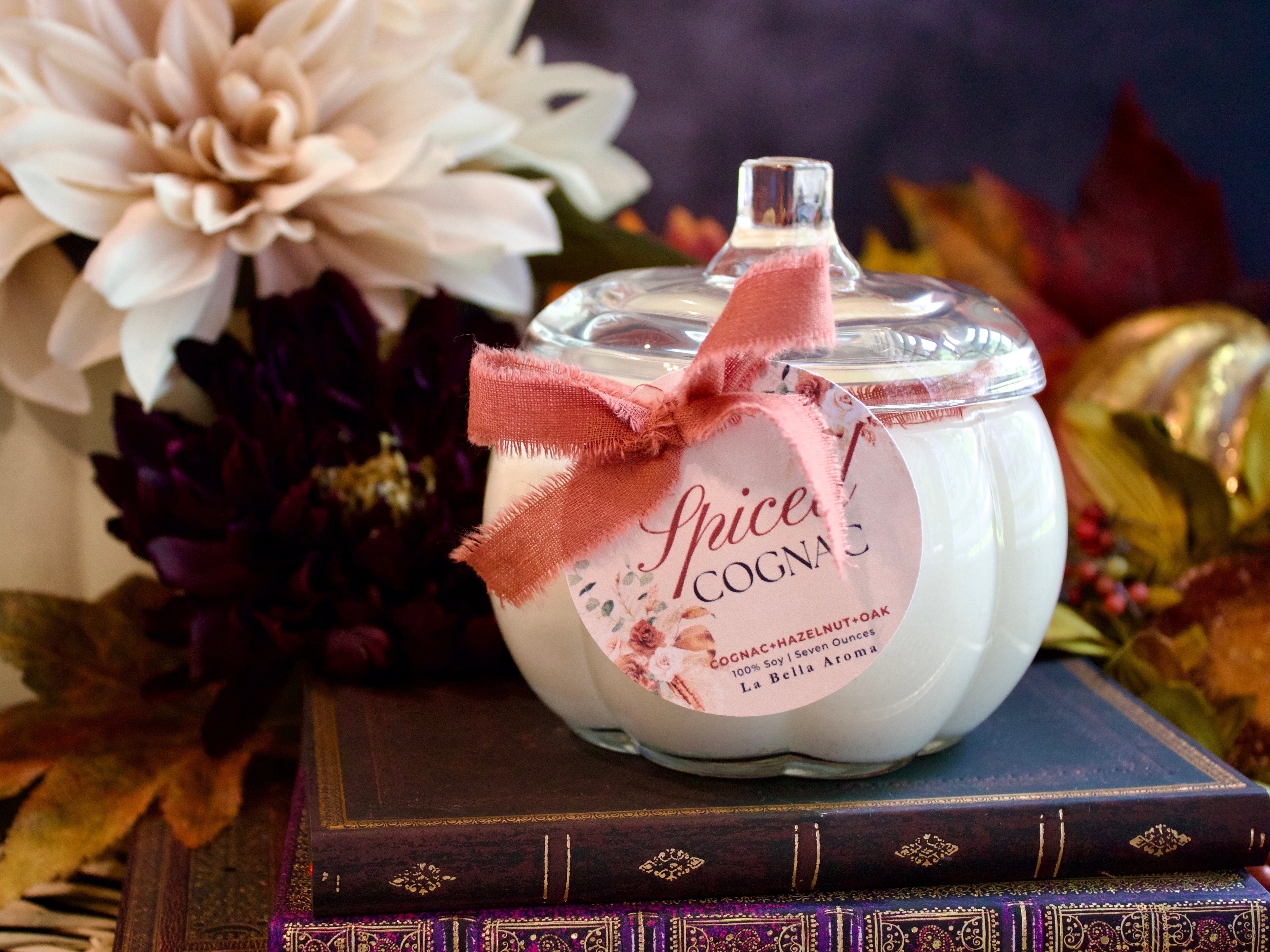 Candle labeled 'Spiced Cognac' with a pink bow on a stack of books with flowers in the background